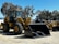 A large yellow construction vehicle with massive tires and a front loader attachment is parked on a paved surface. Several similar machinery units are aligned in the background under a clear blue sky. Tall trees with dense foliage surround the area.