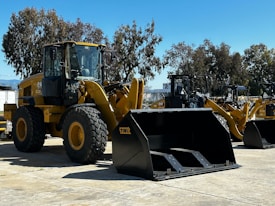 A large yellow construction vehicle with massive tires and a front loader attachment is parked on a paved surface. Several similar machinery units are aligned in the background under a clear blue sky. Tall trees with dense foliage surround the area.
