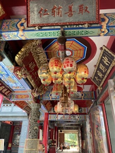 Intricately decorated interior space with vibrant red pillars and ornate, multicolored ceiling beams. Traditional Chinese lanterns with red tassels hang in the center, surrounded by gold and decorative carvings. Chinese calligraphy adorns various plaques and signs, adding cultural and historical elements.