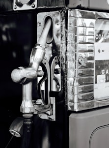 A vintage gas pump with a metallic nozzle and handle attached to it. The structure has a polished chrome finish with visible scratches and wear. Decals and numbers indicating measurements are partially visible on the right side, suggesting its function as a fueling instrument.