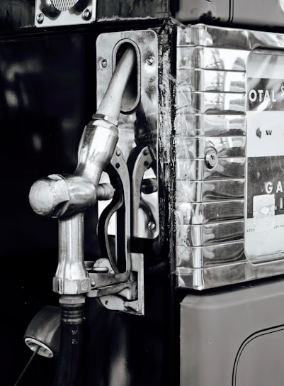 A vintage gas pump with a metallic nozzle and handle attached to it. The structure has a polished chrome finish with visible scratches and wear. Decals and numbers indicating measurements are partially visible on the right side, suggesting its function as a fueling instrument.