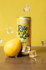 A can of 'Rite n Lite' lemon-flavored drink is placed on a wooden surface with a lemon and several ice cubes surrounding it. The background is a solid yellow, complementing the yellow-themed can and lemon.
