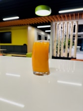A tall glass filled with orange juice sits on a reflective white countertop. The modern interior has warm lighting, with a focus on contemporary design elements like wood paneling and indoor greenery.