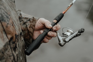 A person wearing a camouflage jacket is holding a fishing rod. The fishing reel is prominently featured, with the hand showing some dirt and wear, suggesting an outdoor activity.