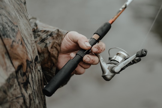 A person wearing a camouflage jacket is holding a fishing rod. The fishing reel is prominently featured, with the hand showing some dirt and wear, suggesting an outdoor activity.