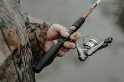 A person wearing a camouflage jacket is holding a fishing rod. The fishing reel is prominently featured, with the hand showing some dirt and wear, suggesting an outdoor activity.