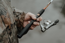 A person wearing a camouflage jacket is holding a fishing rod. The fishing reel is prominently featured, with the hand showing some dirt and wear, suggesting an outdoor activity.