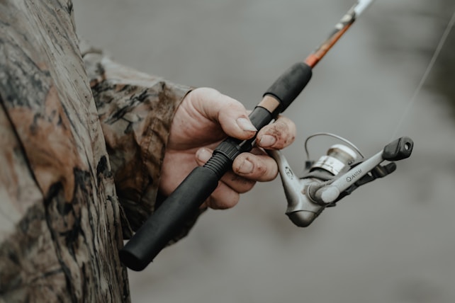A person wearing a camouflage jacket is holding a fishing rod. The fishing reel is prominently featured, with the hand showing some dirt and wear, suggesting an outdoor activity.