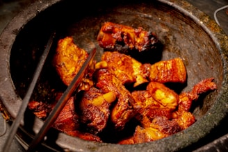 Chunks of cooked meat with a rich, reddish-brown glaze are placed inside a rustic stone vessel. The meat appears juicy and well-cooked, with visible grill marks. A pair of metal tongs rests against the side of the vessel, indicating a barbecue or grilling setting.