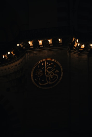 A circular plaque displaying Arabic calligraphy is mounted on a wall. Above, a series of lights on an ornate ledge provide soft illumination, casting a warm glow over the area.
