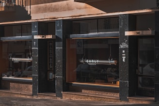 A storefront with large window displays, presumably of a café or bakery named EARLY. The exterior features dark-colored panels and stone finishes, with subtle graffiti on one of the pillars. The windows reflect nearby parked cars and part of the surrounding street scene. Tables and chairs are visible inside, suggesting it is a place for customers to sit and enjoy their purchases.