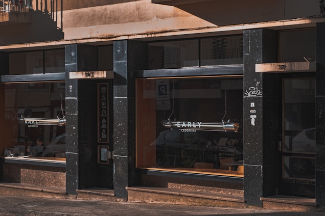 A storefront with large window displays, presumably of a café or bakery named EARLY. The exterior features dark-colored panels and stone finishes, with subtle graffiti on one of the pillars. The windows reflect nearby parked cars and part of the surrounding street scene. Tables and chairs are visible inside, suggesting it is a place for customers to sit and enjoy their purchases.