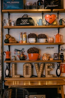 A cozy vintage shelf displays a variety of eclectic items, including retro-style kettles, coffee pots, mugs, and decorative objects like owl figurines and a small statue. On the shelf, an illuminated 'LOVE' sign adds a warm glow to the scene, paired with potted plants and travel souvenirs such as bags with airline logos.