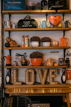 A cozy vintage shelf displays a variety of eclectic items, including retro-style kettles, coffee pots, mugs, and decorative objects like owl figurines and a small statue. On the shelf, an illuminated 'LOVE' sign adds a warm glow to the scene, paired with potted plants and travel souvenirs such as bags with airline logos.