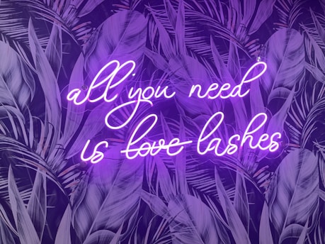 A neon sign with the phrase 'all you need is love lashes' in cursive is displayed against a dark backdrop of stylized palm leaves. The neon light emits a bright purple glow that contrasts with the darker background, creating a vibrant and eye-catching effect.