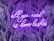 A neon sign with the phrase 'all you need is love lashes' in cursive is displayed against a dark backdrop of stylized palm leaves. The neon light emits a bright purple glow that contrasts with the darker background, creating a vibrant and eye-catching effect.