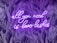 A neon sign with the phrase 'all you need is love lashes' in cursive is displayed against a dark backdrop of stylized palm leaves. The neon light emits a bright purple glow that contrasts with the darker background, creating a vibrant and eye-catching effect.