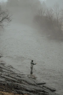 An angler casting a line into a misty river at dawn, surrounded by dense woods.