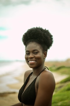 A woman with natural hair styled in a high puff is smiling while standing on a beach. She is wearing a black bikini top. The background features a sandy beach and a blurred horizon with green foliage on the right.