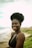A woman with natural hair styled in a high puff is smiling while standing on a beach. She is wearing a black bikini top. The background features a sandy beach and a blurred horizon with green foliage on the right.