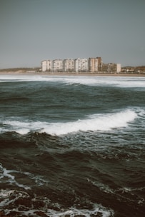 Large ocean waves approach a coastal city with a line of modern high-rise buildings visible in the distance under a cloudy sky. The water appears turbulent, with whitecaps visible on the dark blue sea.