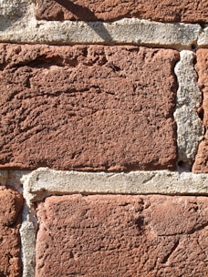 A close-up view of a textured brick wall constructed with red bricks and grey mortar. The bricks have a rough surface and are laid in a horizontal pattern, with visible aggregate in the mortar.
