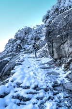 A person wearing a backpack and using trekking poles is hiking up a snow-covered rocky path. The trail is surrounded by snow-laden rocks and bushes, and the sky above is clear with a light blue hue. The scene suggests a cold, winter hike in a mountainous area.