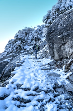 A person wearing a backpack and using trekking poles is hiking up a snow-covered rocky path. The trail is surrounded by snow-laden rocks and bushes, and the sky above is clear with a light blue hue. The scene suggests a cold, winter hike in a mountainous area.