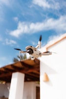 A compact drone with a camera suspended in mid-air against a backdrop of a blue sky with scattered clouds. In the background, a part of a building with white walls and a wooden roof is visible.