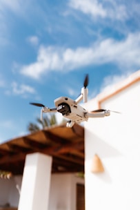 A compact drone with a camera suspended in mid-air against a backdrop of a blue sky with scattered clouds. In the background, a part of a building with white walls and a wooden roof is visible.