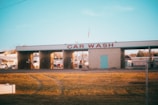 A car wash facility with three empty washing bays, visible under morning or evening light. The building has a sign reading 'CAR WASH' in large red letters. Surrounding the car wash are some parked vehicles and small buildings, and an American flag is seen on top of the structure. The foreground features a stretch of yellowed grass with visible tire tracks.