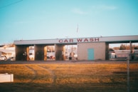 A car wash facility with three empty washing bays, visible under morning or evening light. The building has a sign reading 'CAR WASH' in large red letters. Surrounding the car wash are some parked vehicles and small buildings, and an American flag is seen on top of the structure. The foreground features a stretch of yellowed grass with visible tire tracks.