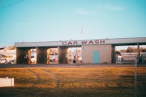 A car wash facility with three empty washing bays, visible under morning or evening light. The building has a sign reading 'CAR WASH' in large red letters. Surrounding the car wash are some parked vehicles and small buildings, and an American flag is seen on top of the structure. The foreground features a stretch of yellowed grass with visible tire tracks.