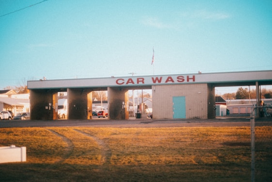 A car wash facility with three empty washing bays, visible under morning or evening light. The building has a sign reading 'CAR WASH' in large red letters. Surrounding the car wash are some parked vehicles and small buildings, and an American flag is seen on top of the structure. The foreground features a stretch of yellowed grass with visible tire tracks.