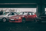 A red car is parked alongside two other vehicles under a dimly lit overpass. The car is a modified Honda with a prominent front bumper and black wheels. People can be seen in the background, partially obscured by one of the other vehicles. The setting appears to be a casual gathering or car meet.