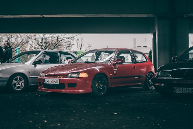 A red car is parked alongside two other vehicles under a dimly lit overpass. The car is a modified Honda with a prominent front bumper and black wheels. People can be seen in the background, partially obscured by one of the other vehicles. The setting appears to be a casual gathering or car meet.