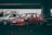 A red car is parked alongside two other vehicles under a dimly lit overpass. The car is a modified Honda with a prominent front bumper and black wheels. People can be seen in the background, partially obscured by one of the other vehicles. The setting appears to be a casual gathering or car meet.
