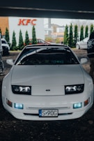 A white sports car is parked in front of a KFC building. The car has a sleek design, with visible headlamps and a polished body. The license plate indicates it's registered in Poland. Two people are partly visible near the car, and the surrounding area has some greenery and commercial buildings in the background.