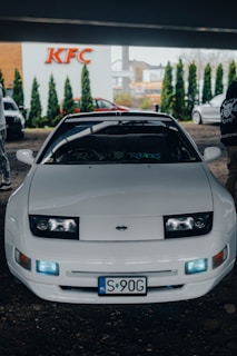 A white sports car is parked in front of a KFC building. The car has a sleek design, with visible headlamps and a polished body. The license plate indicates it's registered in Poland. Two people are partly visible near the car, and the surrounding area has some greenery and commercial buildings in the background.