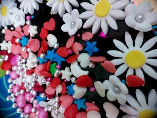 A colorful assortment of various decorative elements including white daisy-like flowers with yellow centers, red hearts, blue stars, pink and red oval shapes, and small silver spheres. These decorations are tightly packed together, creating a vibrant and playful appearance.
