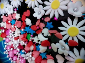 A colorful assortment of various decorative elements including white daisy-like flowers with yellow centers, red hearts, blue stars, pink and red oval shapes, and small silver spheres. These decorations are tightly packed together, creating a vibrant and playful appearance.