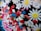 A colorful assortment of various decorative elements including white daisy-like flowers with yellow centers, red hearts, blue stars, pink and red oval shapes, and small silver spheres. These decorations are tightly packed together, creating a vibrant and playful appearance.