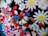 A colorful assortment of various decorative elements including white daisy-like flowers with yellow centers, red hearts, blue stars, pink and red oval shapes, and small silver spheres. These decorations are tightly packed together, creating a vibrant and playful appearance.