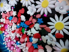A colorful assortment of various decorative elements including white daisy-like flowers with yellow centers, red hearts, blue stars, pink and red oval shapes, and small silver spheres. These decorations are tightly packed together, creating a vibrant and playful appearance.