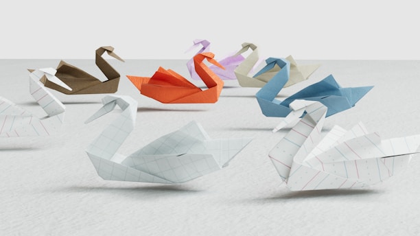 A group of origami swans crafted from various colored and patterned papers, arranged on a light surface. The swans are made from sheets with different textures, including grids, plain colors, and stripes, showing a variety of hues such as blue, orange, brown, and white.