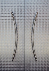 Two vertical metallic handles are mounted on a textured metallic surface, with an abstract geometric pattern. The handles have a sleek and modern design with a slight curve.