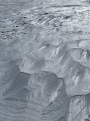 The image captures a detailed view of fine sand or silt with intricate, wavy patterns and ridges sculpted across its surface. The texture is smooth and uniform, with a metallic sheen that glistens in the light, giving the appearance of a serene, windswept landscape.