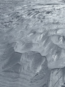 The image captures a detailed view of fine sand or silt with intricate, wavy patterns and ridges sculpted across its surface. The texture is smooth and uniform, with a metallic sheen that glistens in the light, giving the appearance of a serene, windswept landscape.