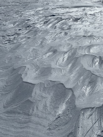 The image captures a detailed view of fine sand or silt with intricate, wavy patterns and ridges sculpted across its surface. The texture is smooth and uniform, with a metallic sheen that glistens in the light, giving the appearance of a serene, windswept landscape.