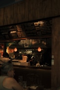 A dimly lit restaurant scene with a focus on a menu board displaying various food items and prices. The interior features hanging light fixtures with warm lighting, giving the space a cozy ambience. Behind the counter, there are glass racks, cooking utensils, and a couple of staff members engaged in work. A patron is seated at a table in the foreground.